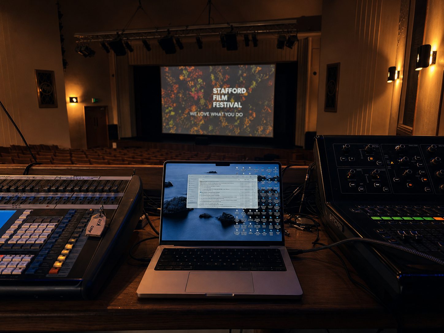 The technical desk at the Mitchell Arts Centre, with a laptop between the lighting and sound consoles and the festival slide on the screen beyond